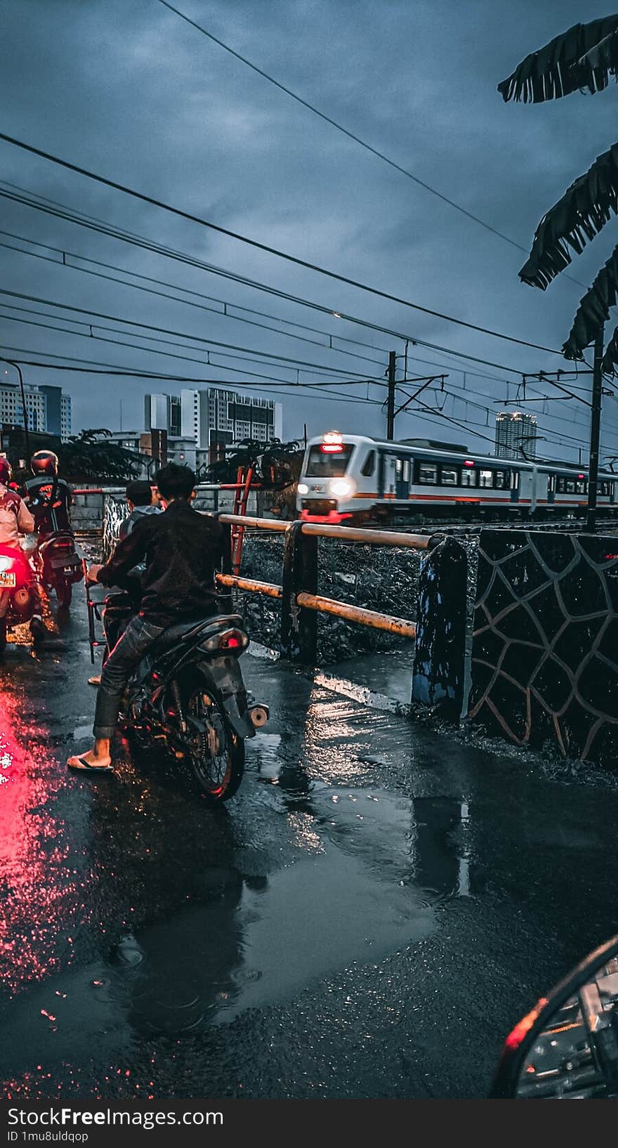 & x22 A wet city street after rain, with motorcyclists waiting at an intersection under cloudy skies. Electric cables are seen crisscrossing above, adding to the atmosphere.