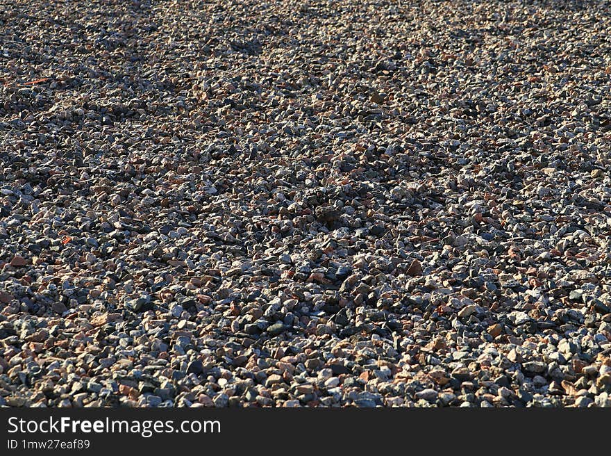 Rubble texture brightly illuminated by morning sunlight. Close-up of small, multicolored pebbles in shades of brown, gray, and red. The lighting creates shadows and highlights, adding depth to the textured surface.