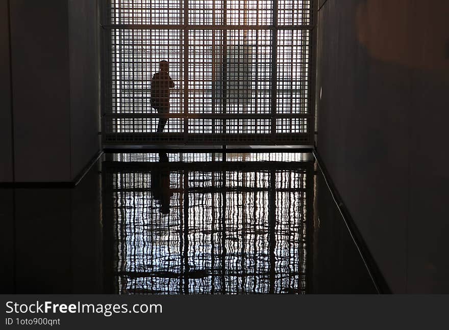 Solitary figure is silhouetted against a geometric patterned glass wall in the dark, reflected perfectly on the glossy floor