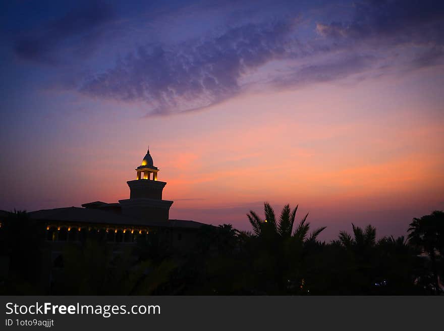 A stunning tower-like building is illuminated against a vibrant sunset sky. Palm trees in the foreground add a tropical touch