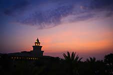 A Stunning Tower-like Building Is Illuminated Against A Vibrant Sunset Sky. Palm Trees In The Foreground Add A Tropical Touch Stock Photos