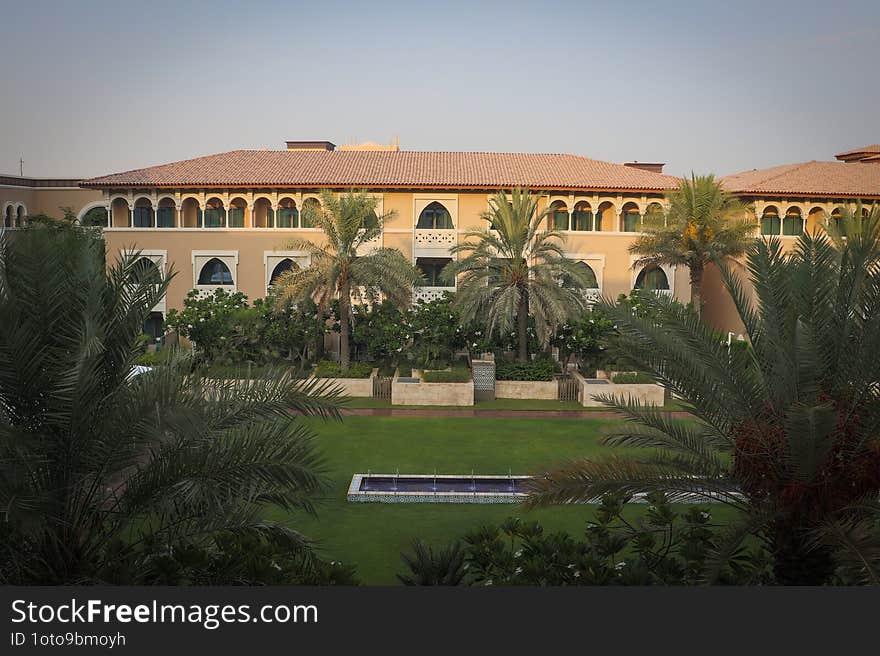 A grand building with arched windows and a red-tiled roof stands majestically. The front garden, adorned with palm trees and a rectangular water feature, adds a touch of serene luxury to the scene.