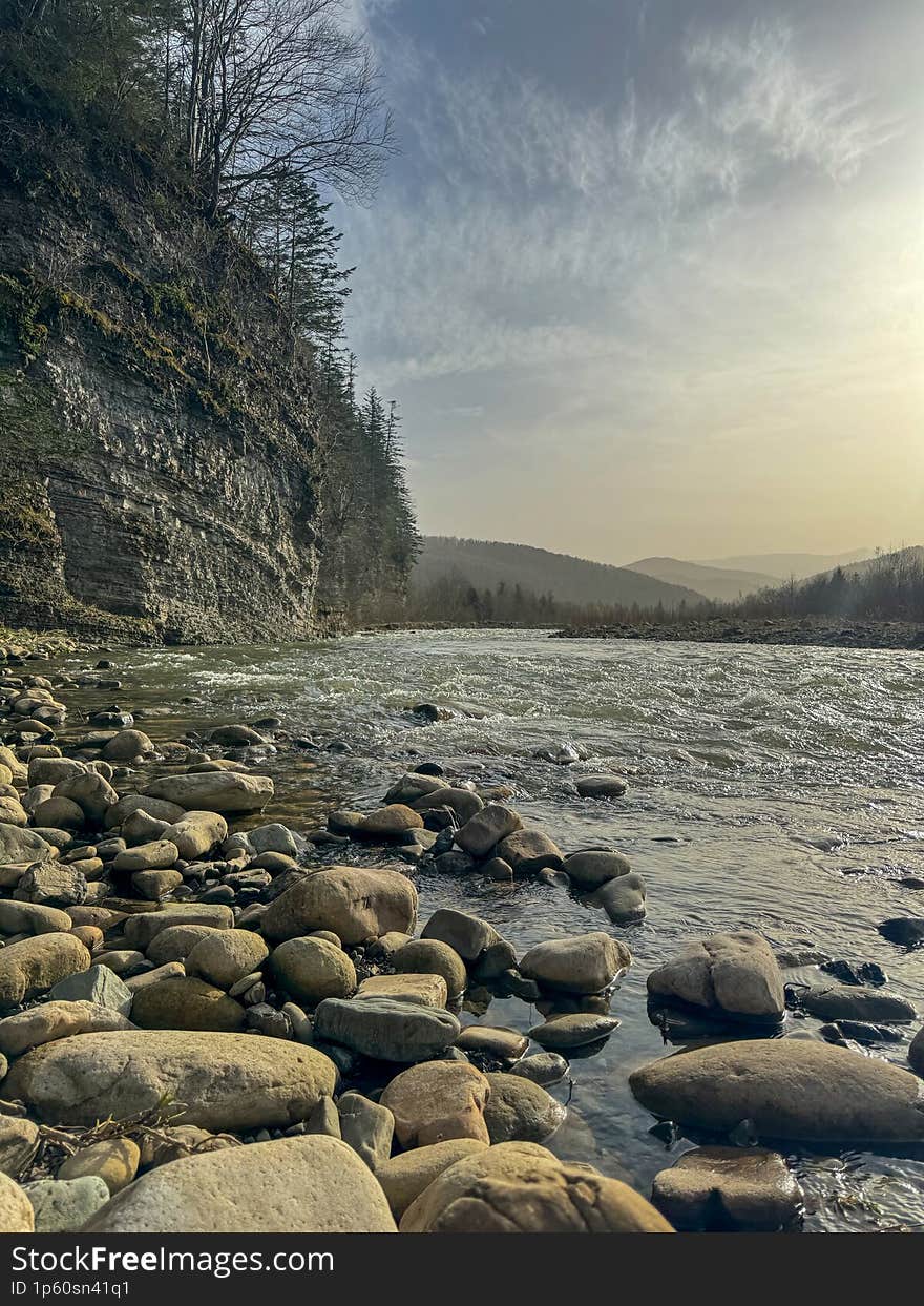 powerful mountain river rushes swiftly past a jagged cliff, its waters churning and foaming as it navigates the rocky terrain. The sun shines brightly above, casting long shadows on the cliff face and illuminating the vibrant green foliage that clings to its sides. The air is filled with the sound of rushing water and the chirping of birds, creating a symphony of nature.