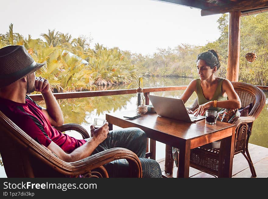 Close up hipster digital nomad couple sit in cafe using laptop work online by scenic river and palm trees in southeast asia