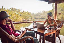 Close Up Hipster Digital Nomad Couple Sit In Cafe Using Laptop Work Online By Scenic River And Palm Trees In Southeast Asia Stock Photography