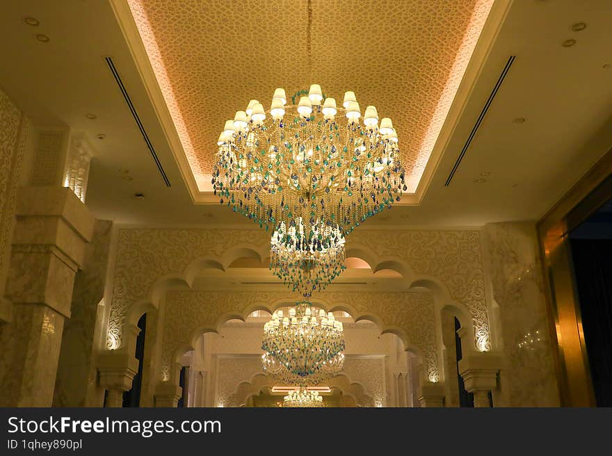 Grand chandelier with multiple tiers of lights and crystals hangs from a ceiling adorned with intricate patterns