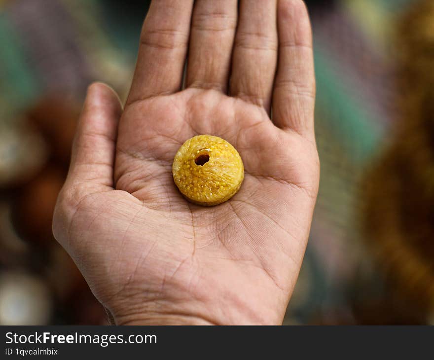 A close-up of a dried fig resting in the palm of a hand. The fig is a deep golden brown color with a slightly wrinkled surface. Th