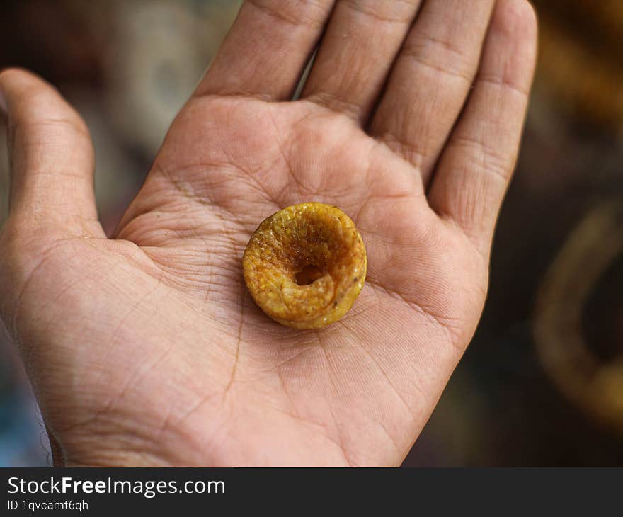 A close-up of a dried fig resting in the palm of a hand. The fig is a deep golden brown color with a slightly wrinkled surface. Th