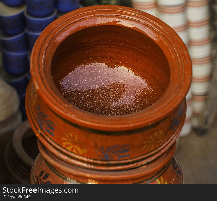 A close-up of a traditional, hand-painted clay pot. The pot is a deep reddish-brown color with intricate designs in blue and yello