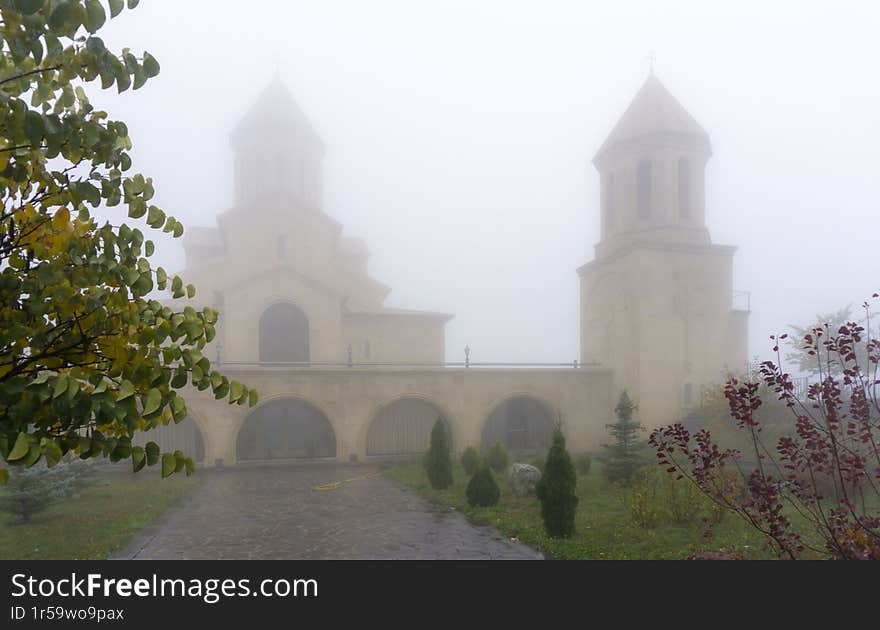 Church yard with grass, bushes and trees, tiled path, bell tower and church in the fog