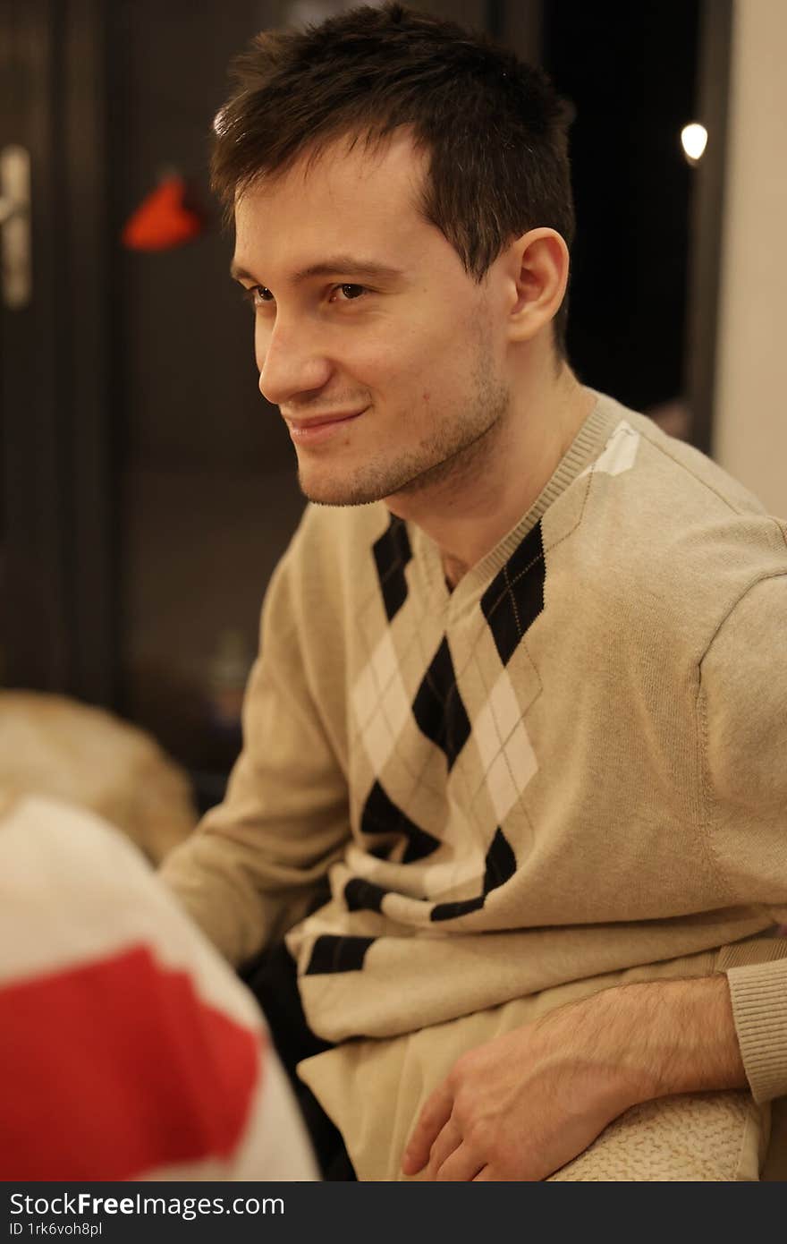 A young man with short dark hair sits in a dimly lit room, wearing a beige sweater with an argyle pattern