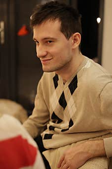 A Young Man With Short Dark Hair Sits In A Dimly Lit Room, Wearing A Beige Sweater With An Argyle Pattern Stock Photo