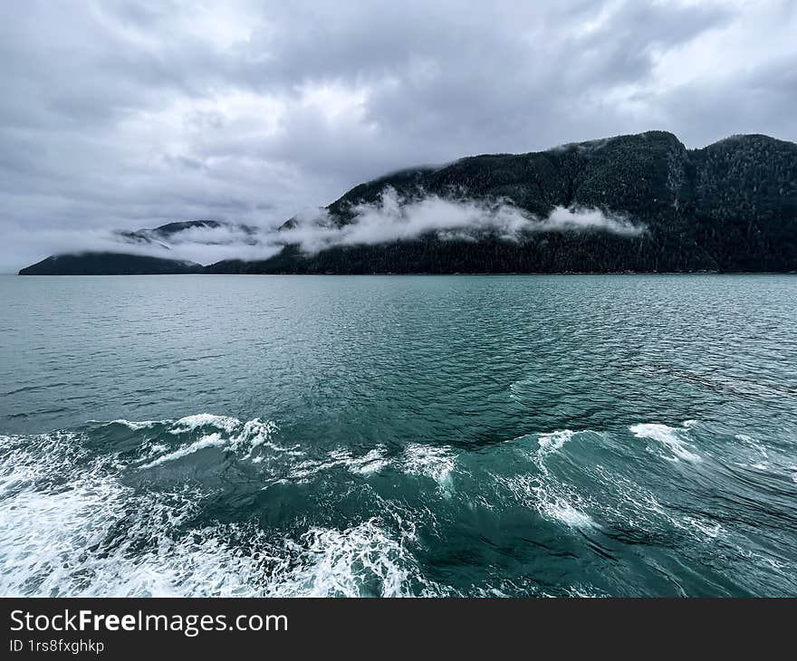 A breathtaking view of the Alaskan wilderness, featuring deep turquoise ocean waters with waves rippling in the foreground and mis