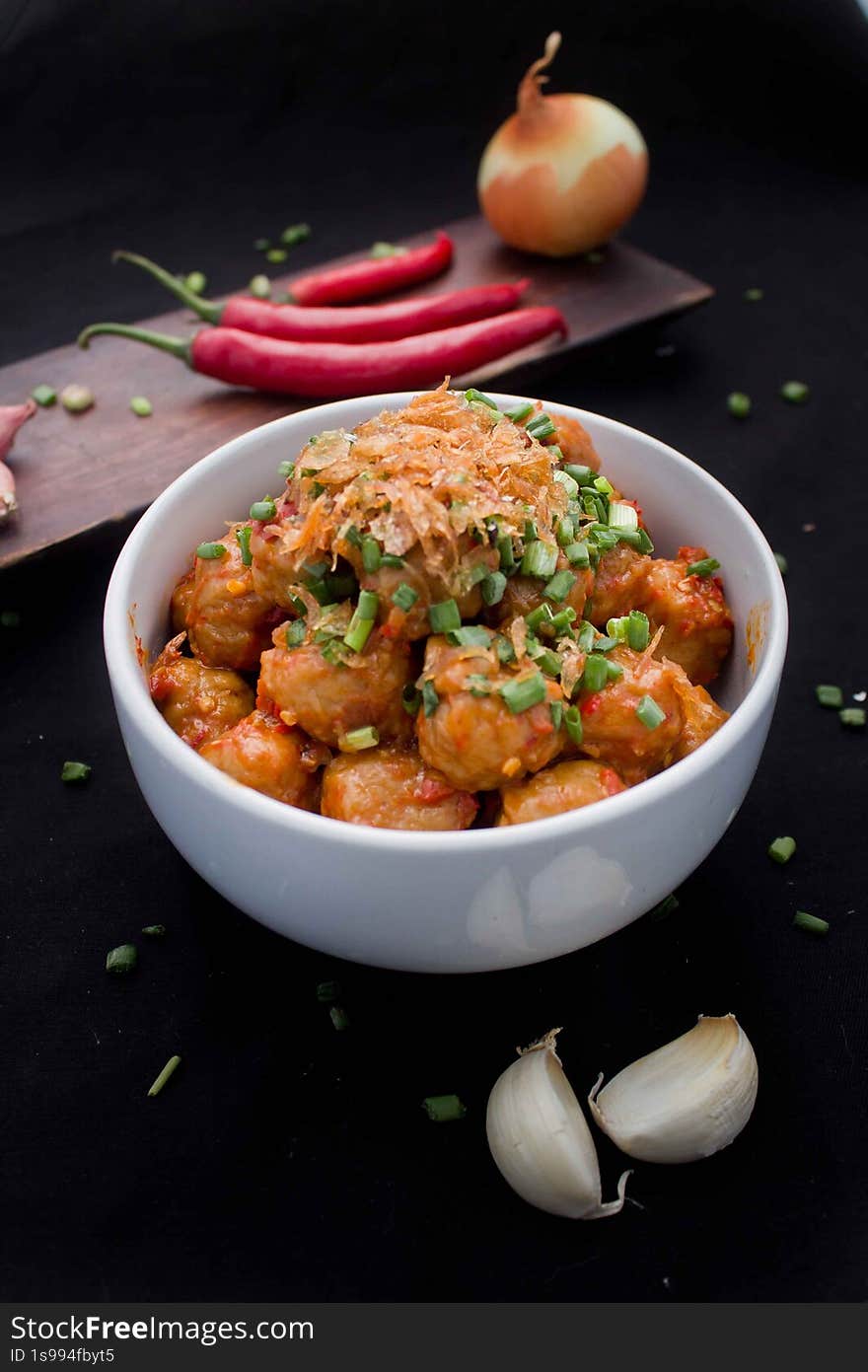the image showcases a bowl of meatballs coated in a vibrant red spicy sauce, generously topped with fried shallots and chopped green onions. the meatballs are served in a white bowl, placed against a black background. in the background, there's a wooden board with red chilies and an onion. two cloves of garlic are placed near the bowl, enhancing the food photography. the image showcases a bowl of meatballs coated in a vibrant red spicy sauce, generously topped with fried shallots and chopped green onions. the meatballs are served in a white bowl, placed against a black background. in the background, there's a wooden board with red chilies and an onion. two cloves of garlic are placed near the bowl, enhancing the food photography.