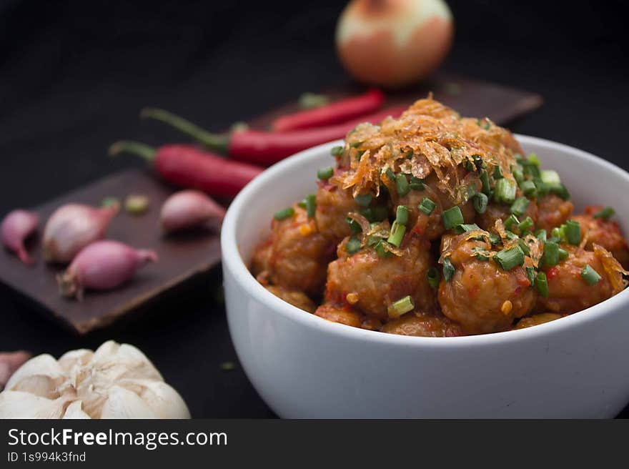 this image showcases a bowl of meatballs coated in a vibrant red chili sauce, generously topped with crispy fried onions and fresh chopped scallions. the meatballs are arranged neatly in a white bowl, which is placed on a black surface. around the bowl, there are cloves of garlic, red chilies, and an onion, suggesting the ingredients used in the dish. the dark background enhances the colors of the food, making it appear appetizing. this image showcases a bowl of meatballs coated in a vibrant red chili sauce, generously topped with crispy fried onions and fresh chopped scallions. the meatballs are arranged neatly in a white bowl, which is placed on a black surface. around the bowl, there are cloves of garlic, red chilies, and an onion, suggesting the ingredients used in the dish. the dark background enhances the colors of the food, making it appear appetizing.