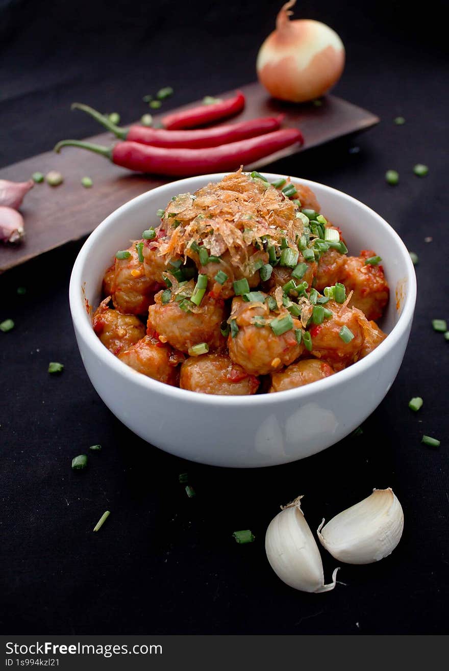 the image showcases a bowl of delicious-looking meatballs coated in a vibrant red chili sauce. the meatballs are generously topped with crispy fried onions and fresh chopped green onions, adding both texture and flavor. in the background, there's a wooden board with red chilies, garlic cloves, and a whole onion, suggesting the ingredients used to prepare the dish. the overall presentation is appetizing and highlights the spicy and savory nature of the meal. the black background enhances the colors of the food. the image showcases a bowl of delicious-looking meatballs coated in a vibrant red chili sauce. the meatballs are generously topped with crispy fried onions and fresh chopped green onions, adding both texture and flavor. in the background, there's a wooden board with red chilies, garlic cloves, and a whole onion, suggesting the ingredients used to prepare the dish. the overall presentation is appetizing and highlights the spicy and savory nature of the meal. the black background enhances the colors of the food.