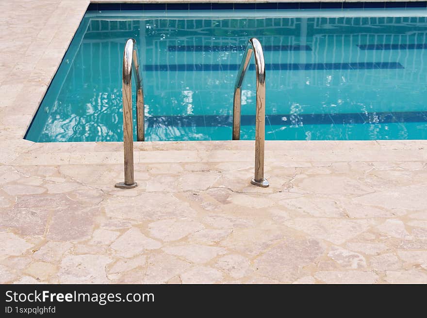 A stainless steel ladder with curved handrails descends into a calm swimming pool with clear blue water. The edge of the pool is s