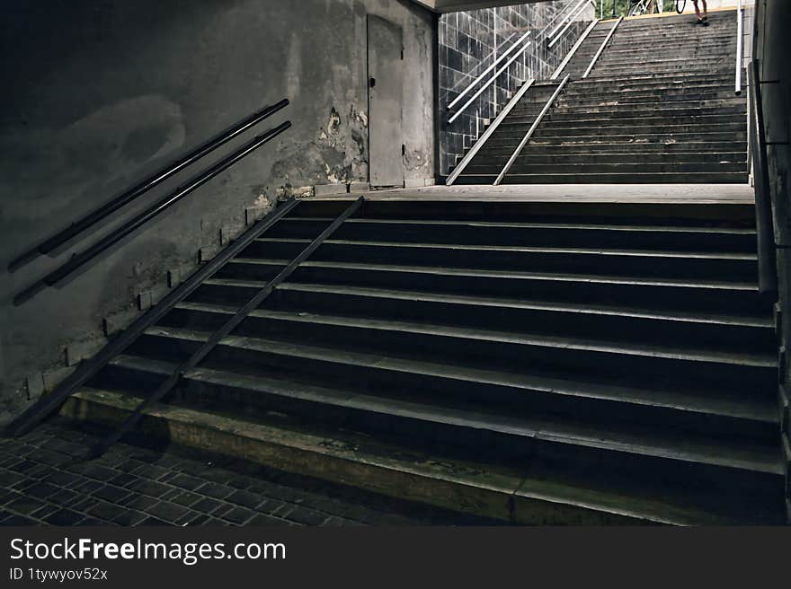 A Dimly Lit Underground Passage With Two Sets Of Stairs Leading Upward. The Worn Walls, Peeling Paint, And Metal Handrails Create