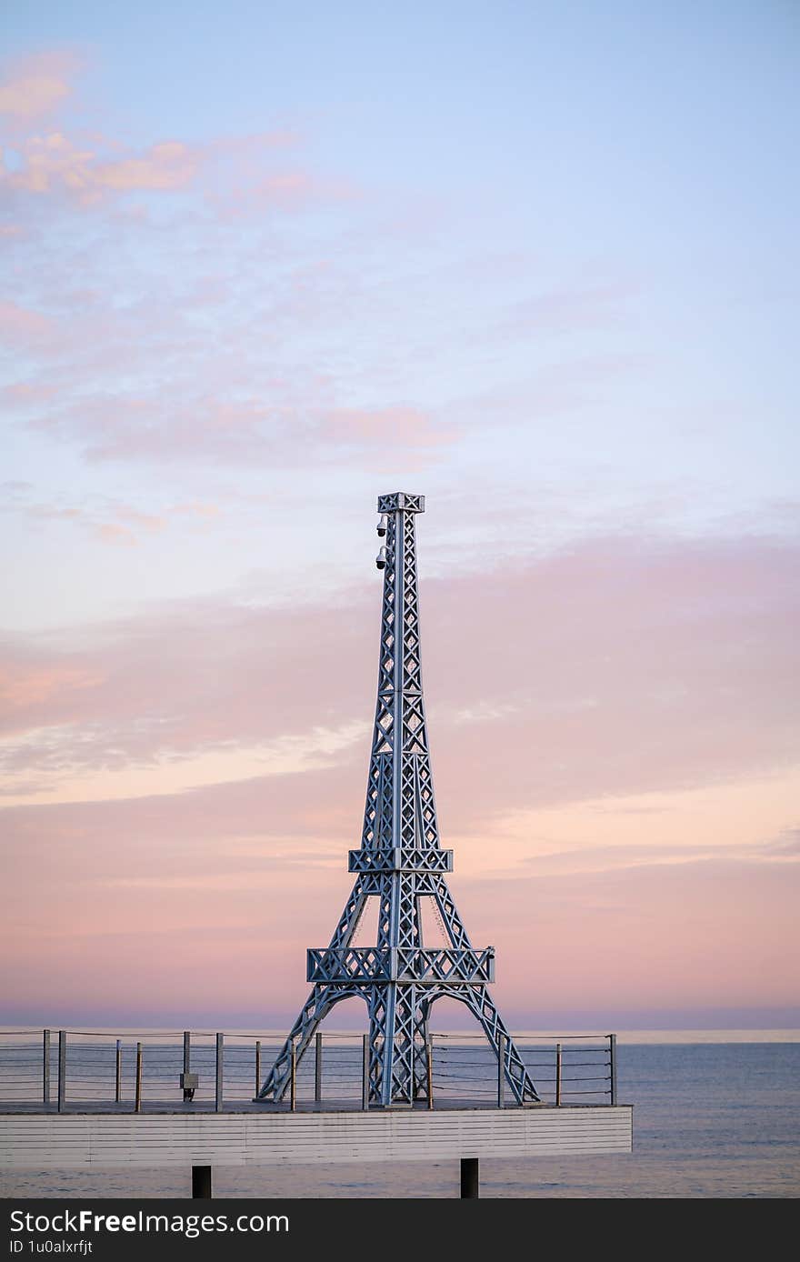 Vivid Eiffel Tower replica on pier at sunset with pastel sky and calm sea, illuminated by soft evening light. No people. Ideal for travel, architecture, scenic imagery.