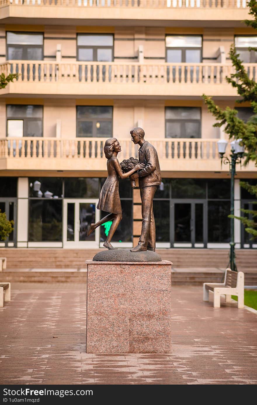 Bronze Statue Of Couple Exchanging Flowers In Front Of Modern Building With Balconies, Featuring Woman In Dress And Man In Casual