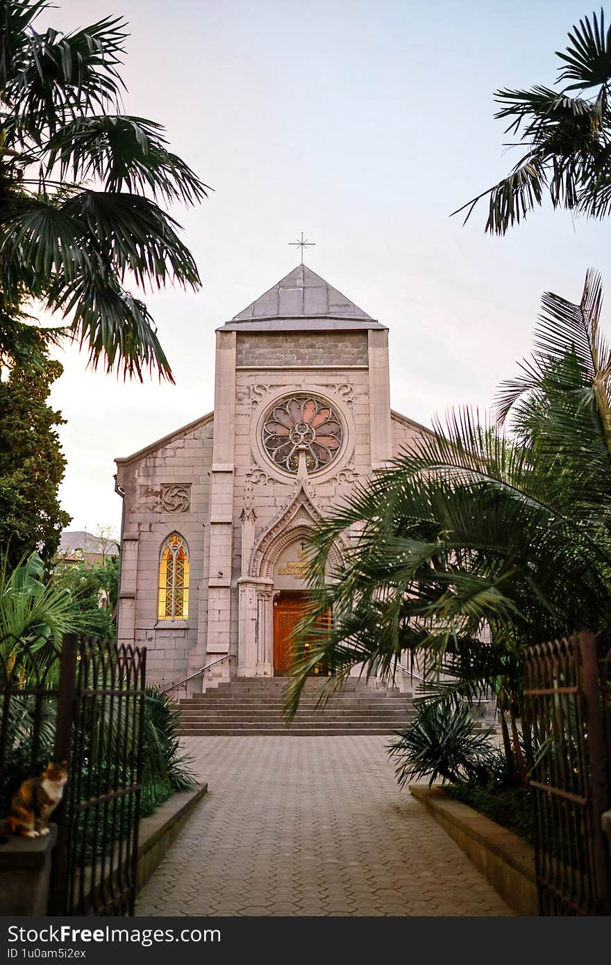 Historic Church Facade With Circular Stained Glass, Surrounded By Palm Trees At Sunset. Wooden Door, Steps And Cat On Fence. Ideal