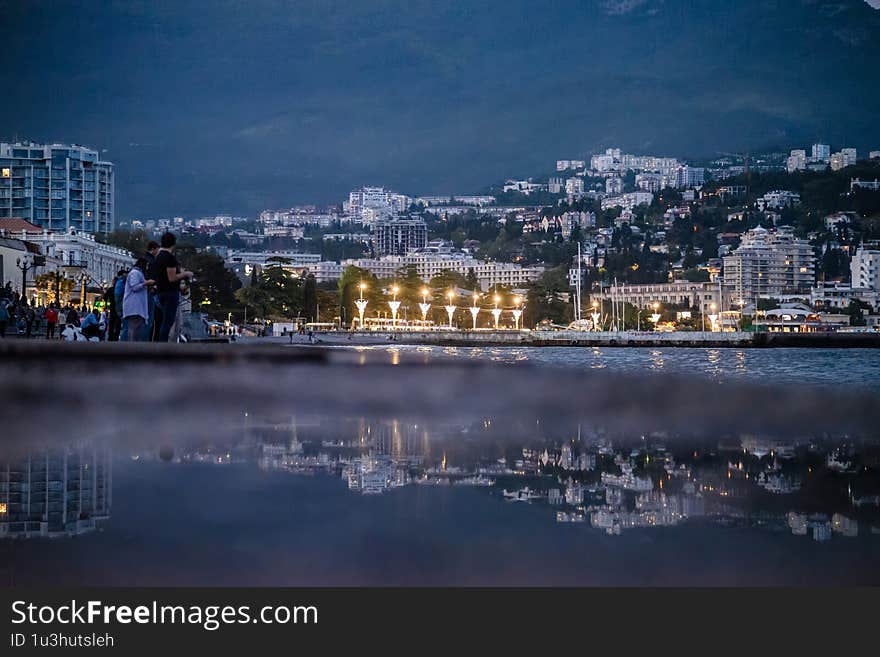 Coastal cityscape at dusk with buildings reflecting on calm water, people on promenade, and hillside in background. Lights glow softly. Perfect for travel, urban, or evening themes