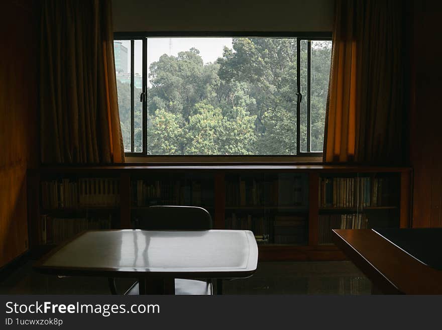 A cozy room features a wooden table, chair, bookshelf, and large curtained window framing serene greenery beyond, evoking calm.