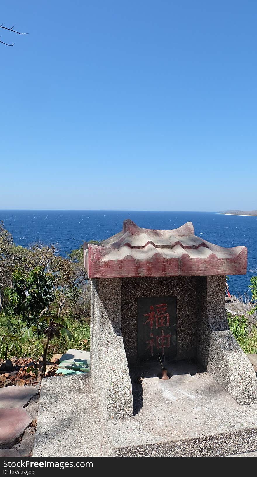A small Asian stone shrine with Chinese characters overlooking the ocean under clear blue sky. Captured in natural daylight, symbolizing peace, spirituality, and coastal culture