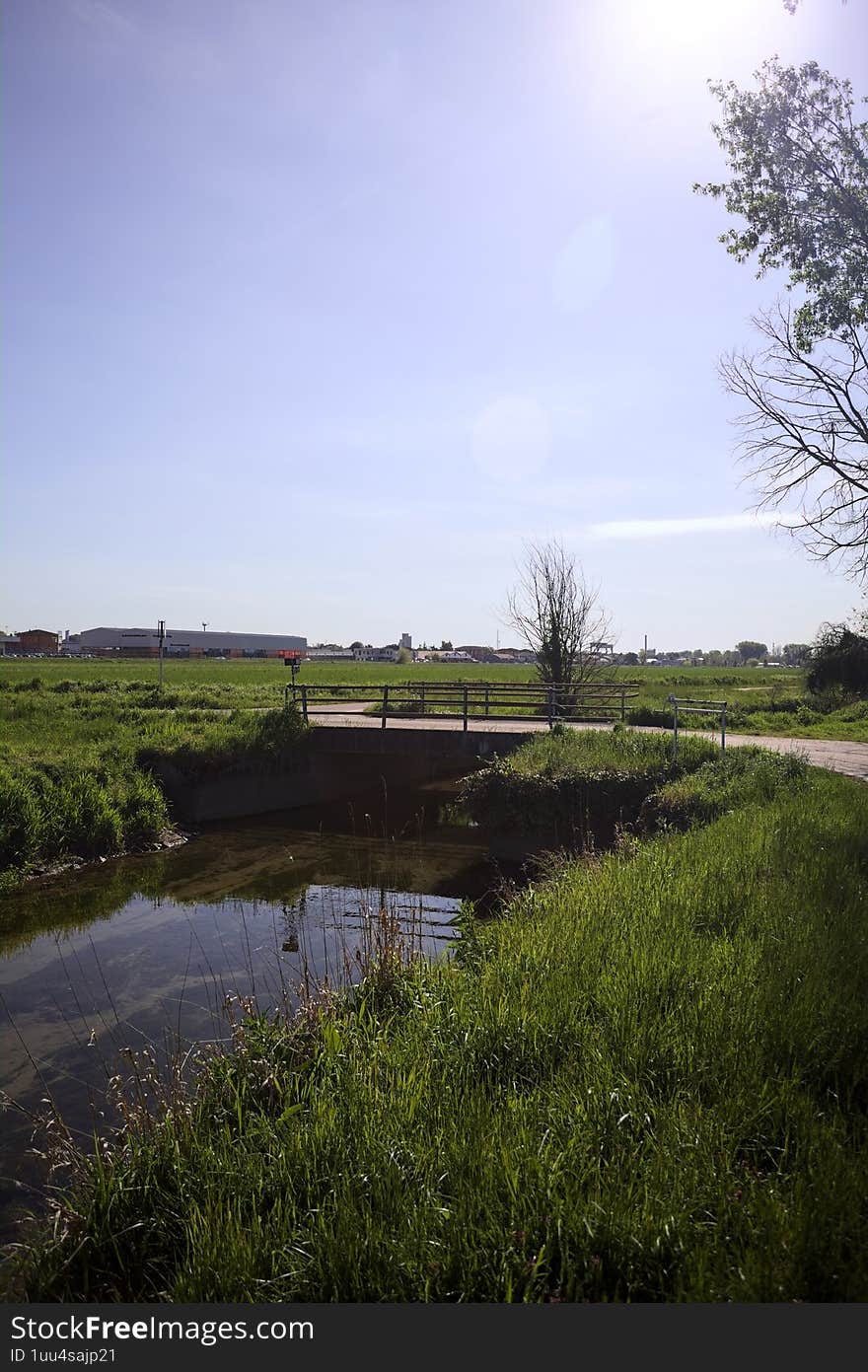 Trail next to a stream of water with a small concrete bridge passing over it and a grass field on a sunny day in the italian countryside. Trail next to a stream of water with a small concrete bridge passing over it and a grass field on a sunny day in the italian countryside