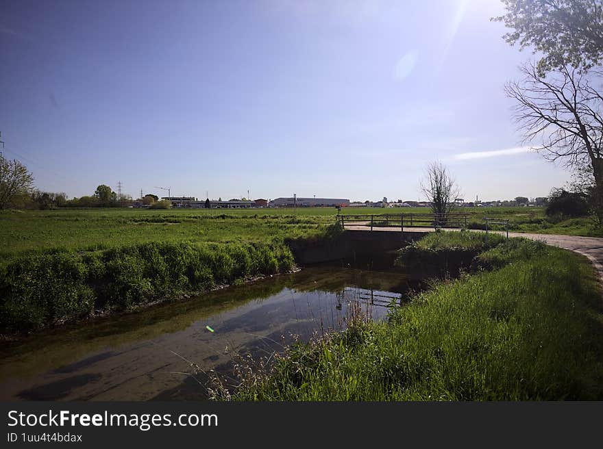 Trail next to a stream of water with a small concrete bridge passing over it and a grass field on a sunny day in the italian countryside. Trail next to a stream of water with a small concrete bridge passing over it and a grass field on a sunny day in the italian countryside