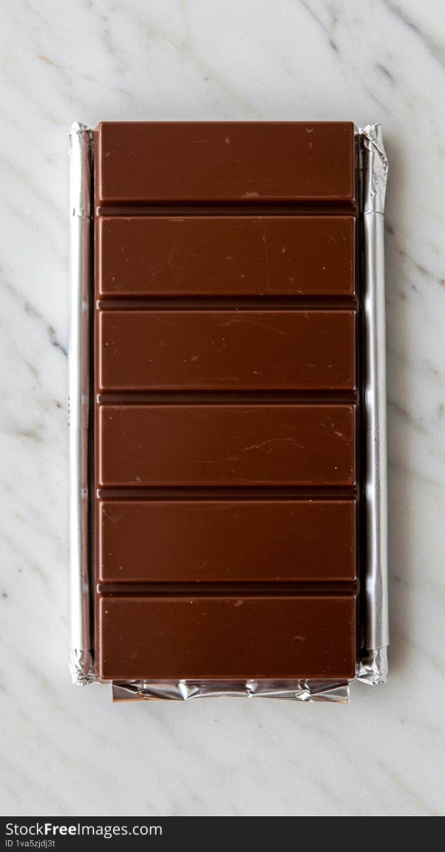 A chocolate bar with a glossy, dark brown surface is divided into uniform rectangular segments. It is partially wrapped in shiny, silver foil, which is slightly crumpled around the edges. The background is a smooth, pale marble surface with subtle gray veining, enhancing the presentation of the chocolate. Each segment is evenly sized, allowing for easy breaking. The bar's simplicity and symmetry highlight its classic design and appeal to chocolate enthusiasts.