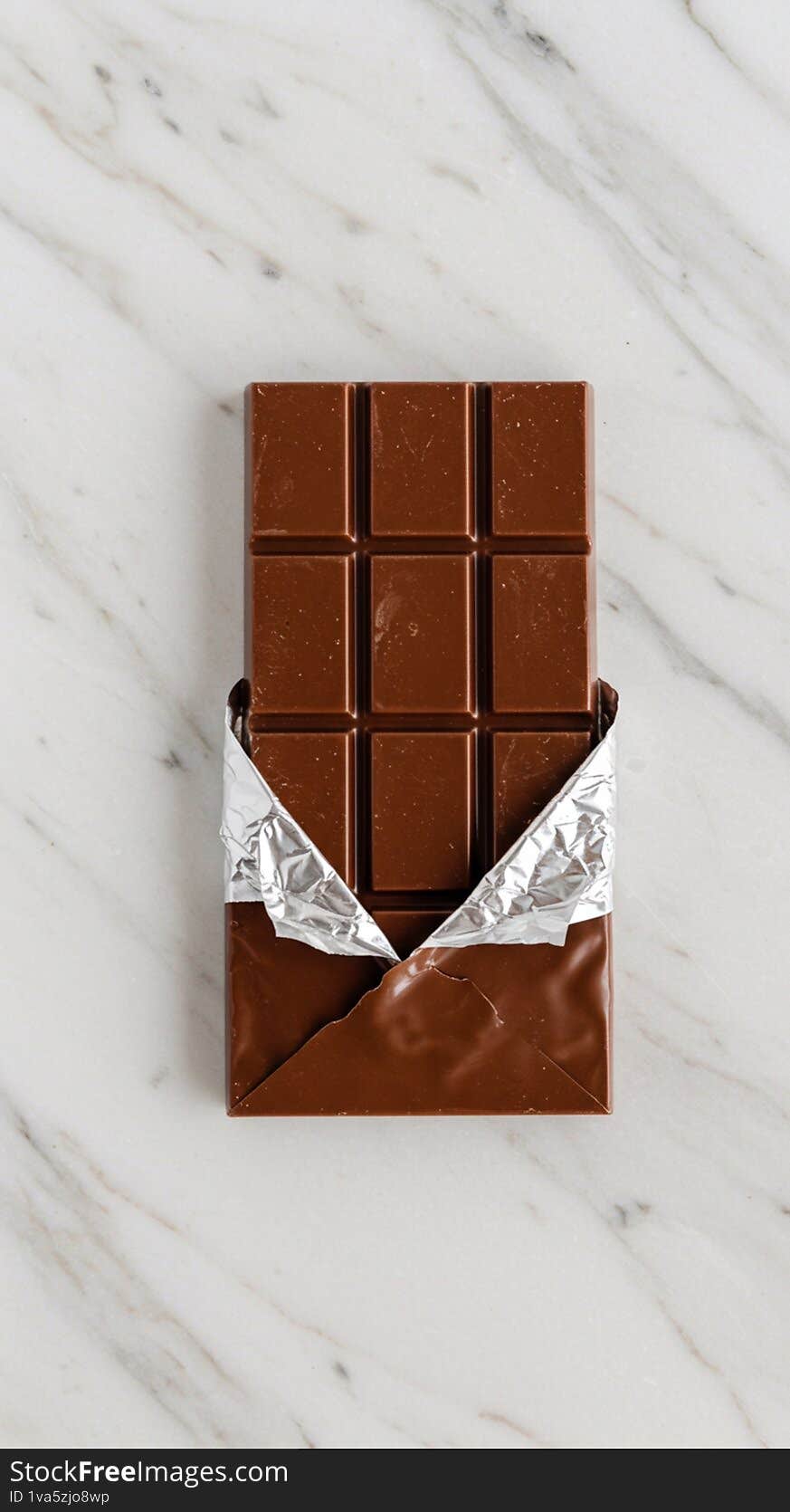 A chocolate bar partially unwrapped, revealing a rich brown surface divided into rectangular segments. The wrapper is a combination of metallic silver foil and brown paper, folded back to expose the chocolate. It rests on a smooth, white marble surface with delicate grey veining.