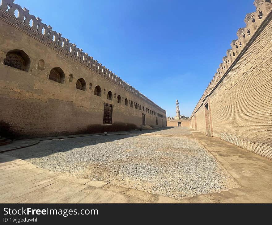 A quiet courtyard surrounded by the historic walls of a centuries-old mosque in Cairo, Egypt. The powerful symmetry, textured stone, and distant minaret evoke the grandeur and serenity of early Islamic architecture. A moment of timeless beauty beneath a clear blue sky.