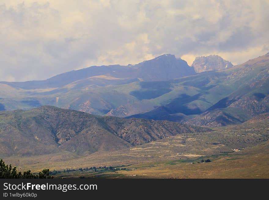 Rolling hills and rugged peaks of the Trans-Ili Alatau Mountains stretch across the horizon under a cloud-filled sky. The mountains display varied hues of green and brown, indicating diverse vegetation and terrain. Below, a spacious, verdant valley unfolds, dotted with patches of lush greenery and sparse tree clusters. The scene captures the majestic and expansive beauty of this mountainous landscape, typical of the Almaty region in Kazakhstan, offering a serene and natural panorama. Rolling hills and rugged peaks of the Trans-Ili Alatau Mountains stretch across the horizon under a cloud-filled sky. The mountains display varied hues of green and brown, indicating diverse vegetation and terrain. Below, a spacious, verdant valley unfolds, dotted with patches of lush greenery and sparse tree clusters. The scene captures the majestic and expansive beauty of this mountainous landscape, typical of the Almaty region in Kazakhstan, offering a serene and natural panorama.