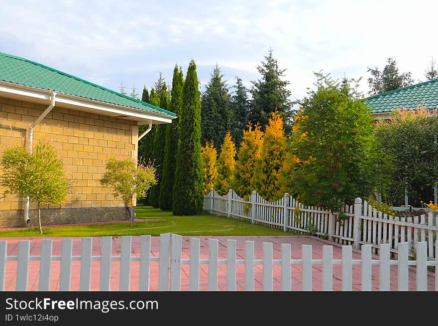Sunlit Garden with Two Yellow-Brick Buildings, Green Roofs, and White Picket Fence, Framed by Lush Trees