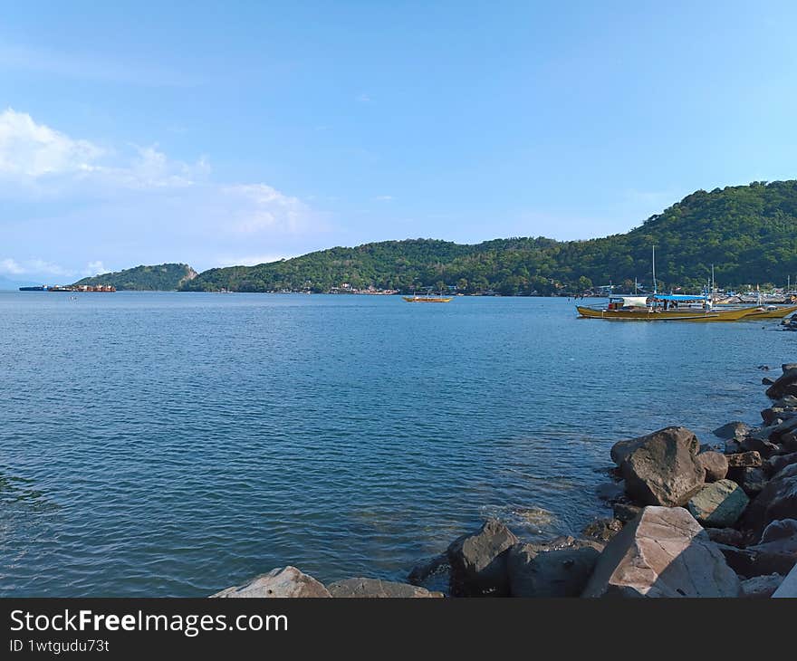 A lively and heartwarming moment captured by the sea. The photo shows a quiet coastal view with small fishing boats gently floating near the shore, their weathered colors adding charm to the scene.