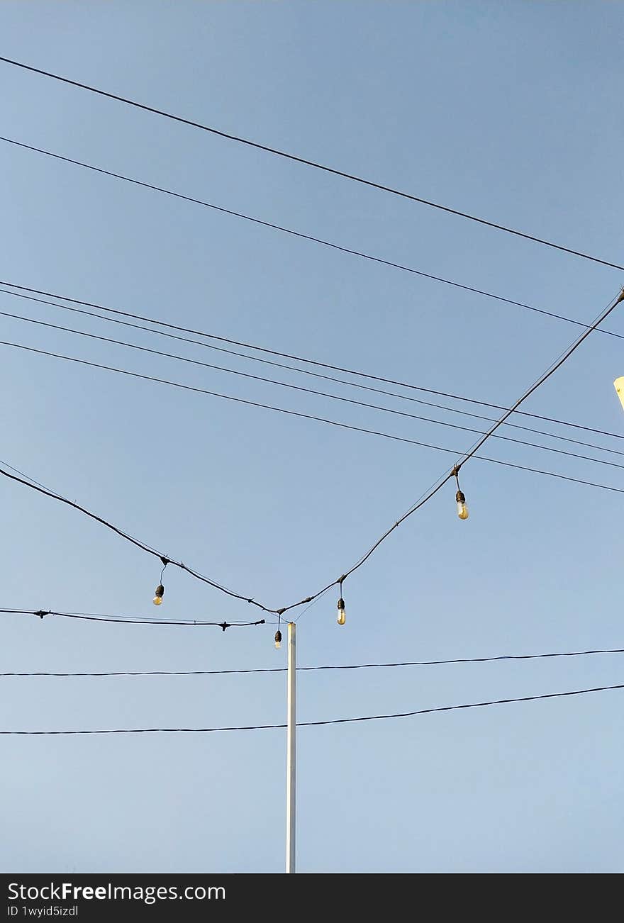 A serene blue sky serves as the backdrop for a series of hanging light bulbs strung across power lines, creating a charming and in