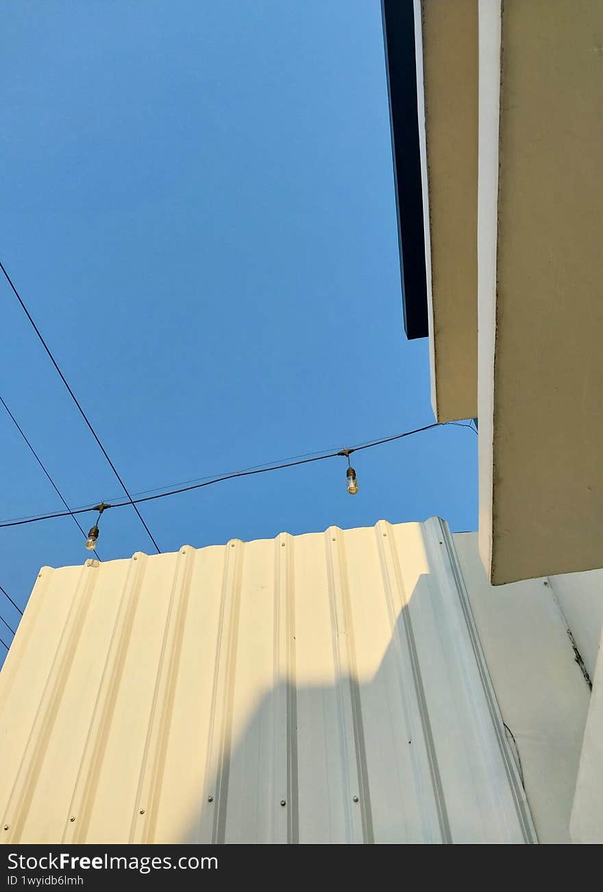 Minimalist outdoor scene featuring a clear blue sky, string lights, a modern beige wall, and part of a building with clean lines, creating a calm and contemporary atmosphere
