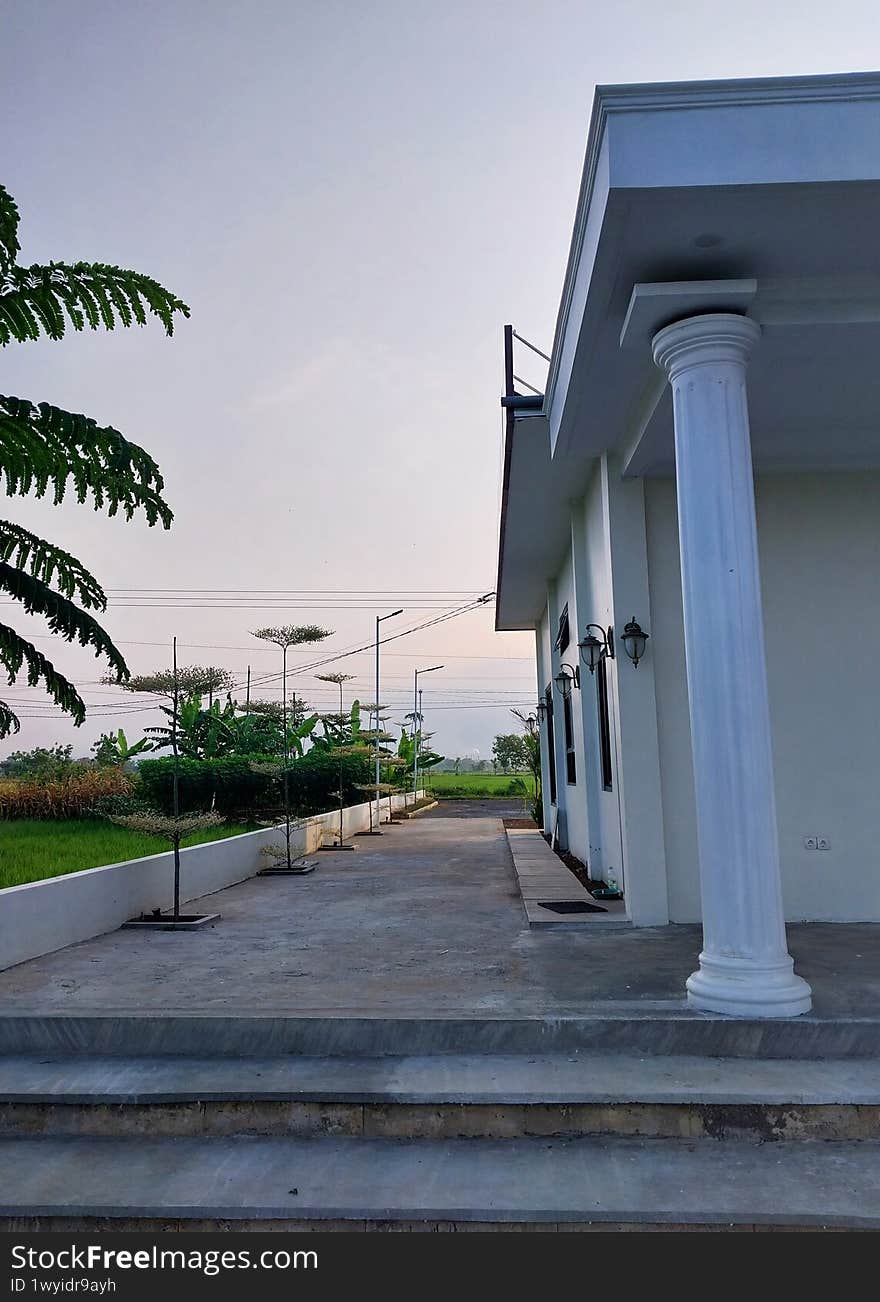 A modern house exterior featuring elegant columns, a spacious entrance, and lush greenery. The serene landscape and clear sky create a peaceful atmosphere, perfect for family living