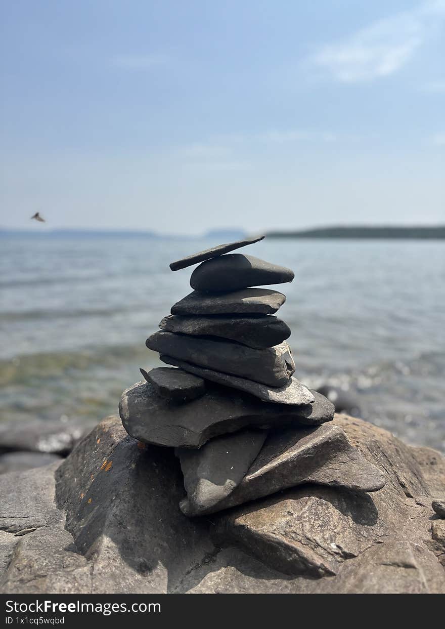 The image captures a serene scene on the shore of Lake Superior, where an intricate pile of rocks, carefully stacked by nature or human hand, rests amidst the tranquil beauty of the landscape. The rocks form a small, yet striking cairn, their varied shapes and sizes creating an almost harmonious balance as they rise against the backdrop of the vast, calm waters. In the distance, the crystal-clear lake stretches out toward the horizon, where the sky meets the water in a gentle gradient of blues and soft cloud formations. The rocky shoreline, covered in smaller stones and pebbles, contrasts with the smoothness of the lake, creating a peaceful juxtaposition between solid earth and fluid water. The setting feels both meditative and timeless, a quiet testament to the power of nature and the art of human expression.