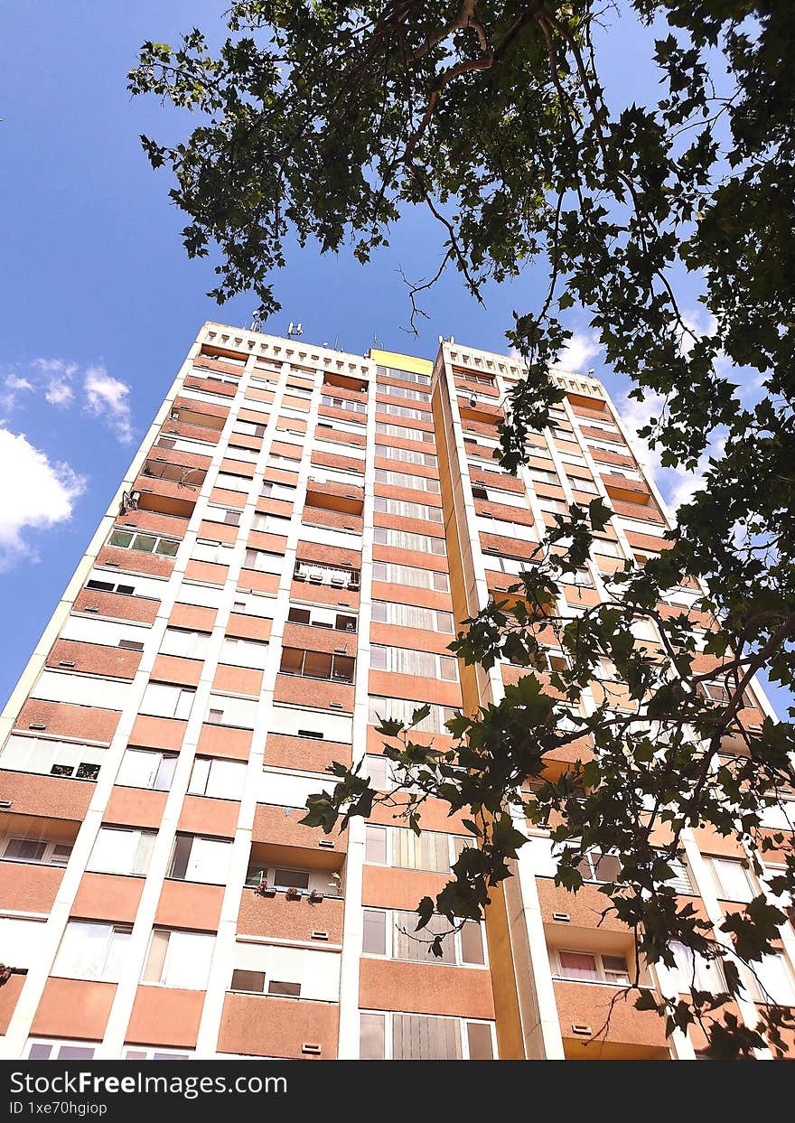 A multi-story panel building, photographed upwards from below, with leafy trees and a blue sky.