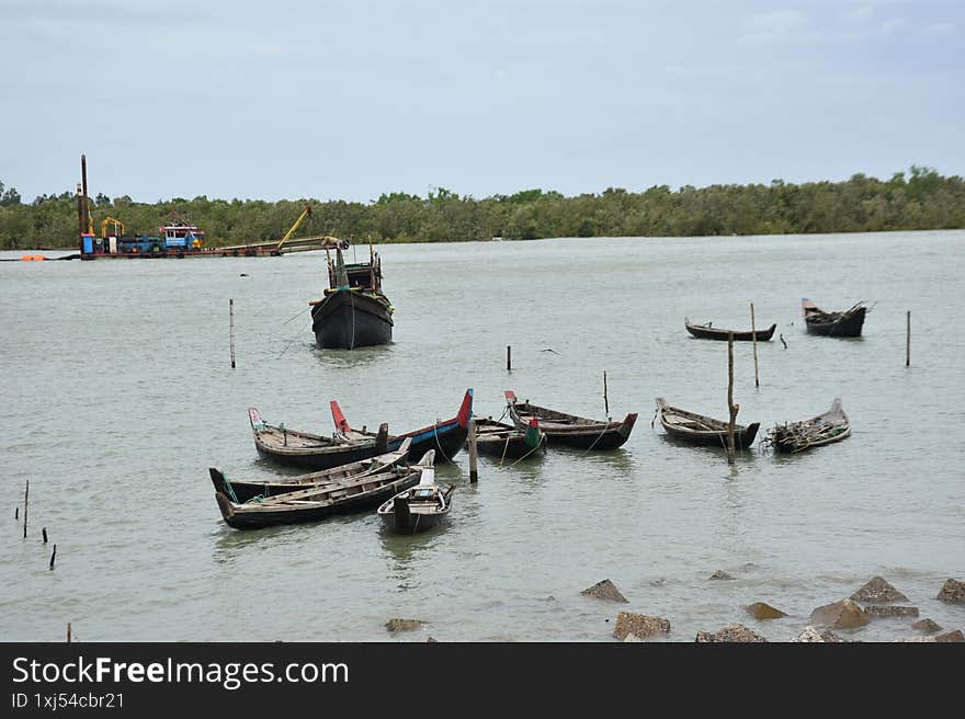 A wide shot of a cluster of traditional wooden boats, some moored to wooden poles, floating on a wide river. In the background, a larger, more rustic boat is visible, with a dense line of trees on the distant bank. The calm, subtle color of the water and the overcast sky create a peaceful atmosphere. This image is ideal for themes of local transport, traditional culture, rivers, and the rural environment in Cox's Bazar, Bangladesh.
