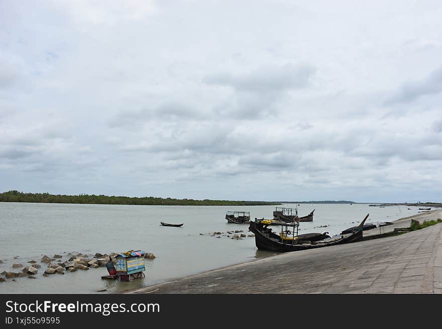 A wide shot of a cluster of traditional wooden boats, some moored to wooden poles, floating on a wide river. In the background, a larger, more rustic boat is visible, with a dense line of trees on the distant bank. The calm, subtle color of the water and the overcast sky create a peaceful atmosphere. This image is ideal for themes of local transport, traditional culture, rivers, and the rural environment in Cox's Bazar, Bangladesh.