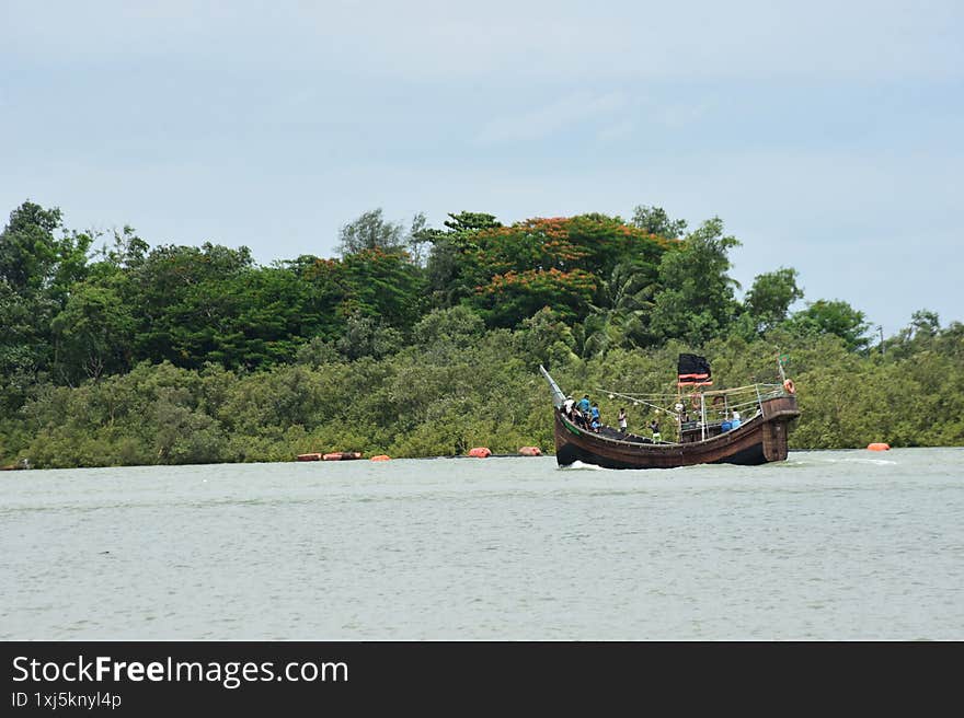 A wide shot of a traditional wooden boat, likely a fishing or transport vessel, navigating a wide river under a hazy sky. The boat is filled with people and equipment, suggesting a journey or daily activity. The river's banks are lined with dense, lush green trees and vegetation, creating a rich natural backdrop. This image captures a moment of local river life and transport in Cox's Bazar, Bangladesh, perfect for themes of travel, culture, rivers, and rural environments. A wide shot of a traditional wooden boat, likely a fishing or transport vessel, navigating a wide river under a hazy sky. The boat is filled with people and equipment, suggesting a journey or daily activity. The river's banks are lined with dense, lush green trees and vegetation, creating a rich natural backdrop. This image captures a moment of local river life and transport in Cox's Bazar, Bangladesh, perfect for themes of travel, culture, rivers, and rural environments