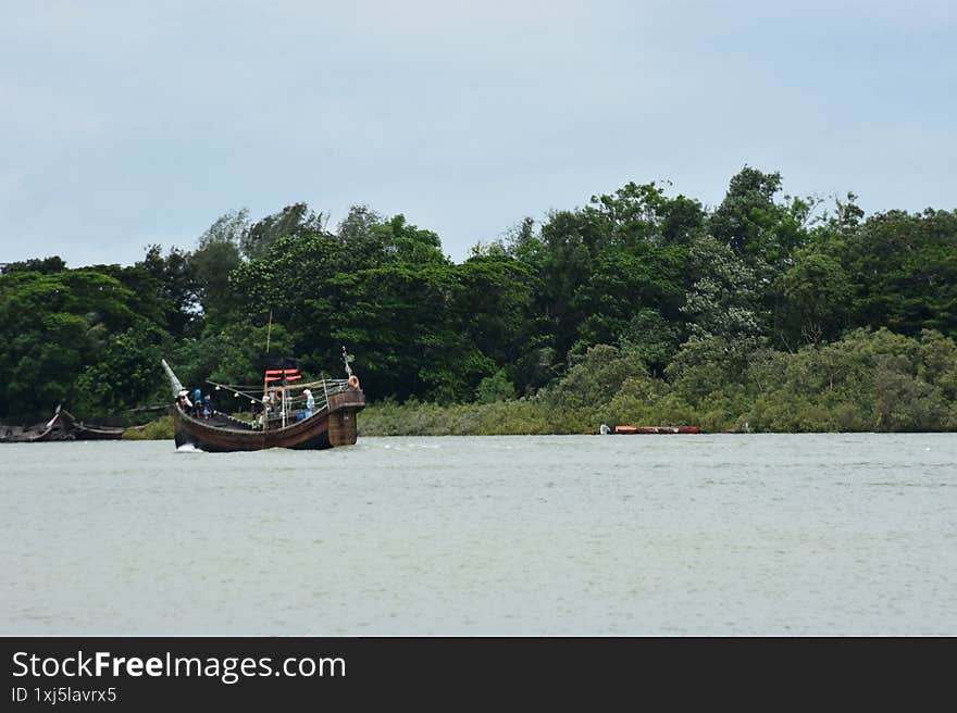 A wide shot of a traditional wooden boat, likely a fishing or transport vessel, navigating a wide river under a hazy sky. The boat is filled with people and equipment, suggesting a journey or daily activity. The river's banks are lined with dense, lush green trees and vegetation, creating a rich natural backdrop. This image captures a moment of local river life and transport in Cox's Bazar, Bangladesh, perfect for themes of travel, culture, rivers, and rural environments. A wide shot of a traditional wooden boat, likely a fishing or transport vessel, navigating a wide river under a hazy sky. The boat is filled with people and equipment, suggesting a journey or daily activity. The river's banks are lined with dense, lush green trees and vegetation, creating a rich natural backdrop. This image captures a moment of local river life and transport in Cox's Bazar, Bangladesh, perfect for themes of travel, culture, rivers, and rural environments