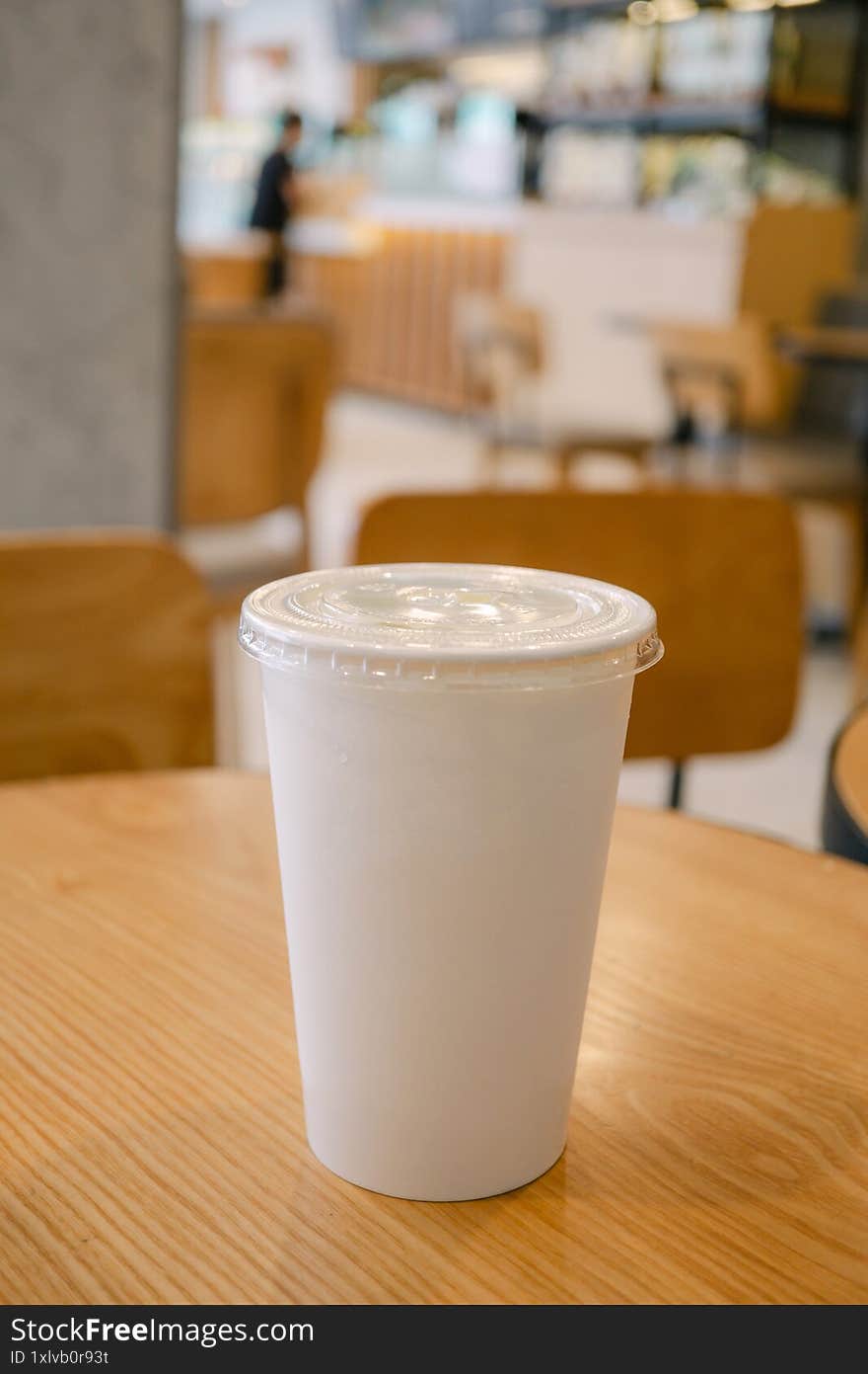 A white plastic cup with a clear lid sits on a wooden table in a cozy caf�, with blurred chairs and soft lighting in the background.