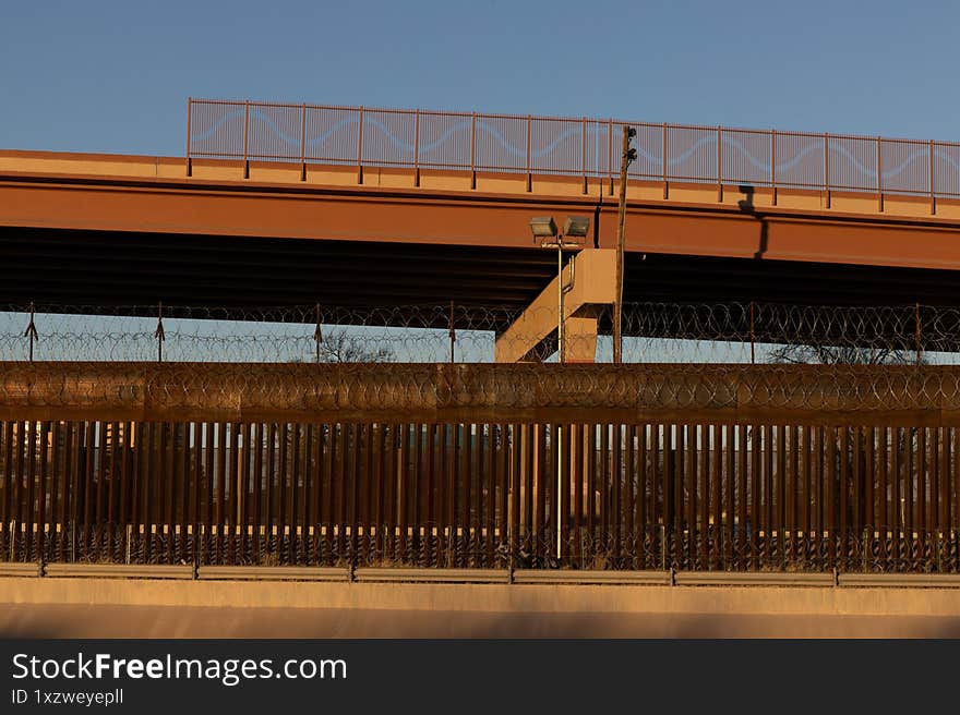 A high metallic barrier with barbed wire marks the tense boundary between Ciudad Juarez and the US under a serene sky