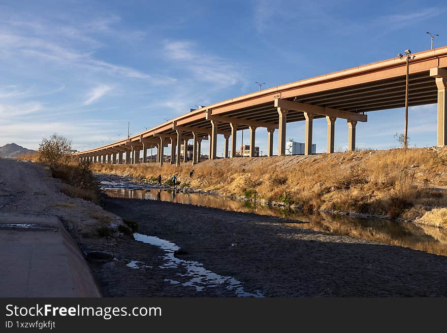 Migrants traverse the Rio Grande under a vast overpass, bordering Ciudad Juarez, amidst sparse flora under a partly cloudy sky