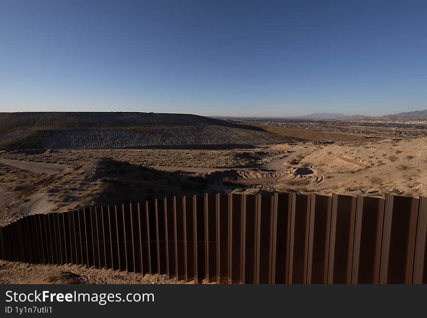 A section of the border barrier in Ciudad Juarez, a vital migration route, stands tall under a clear blue sky, with a dusty trail running alongside
