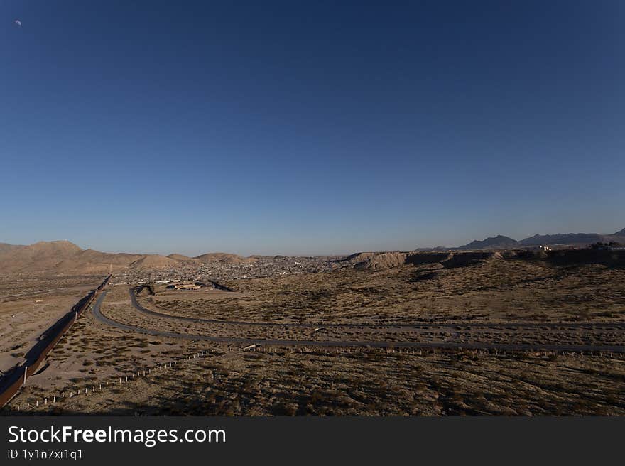 A barrier separating Ciudad Juarez from the US, stretches across a barren landscape, with a road meandering alongside under a clear blue sky